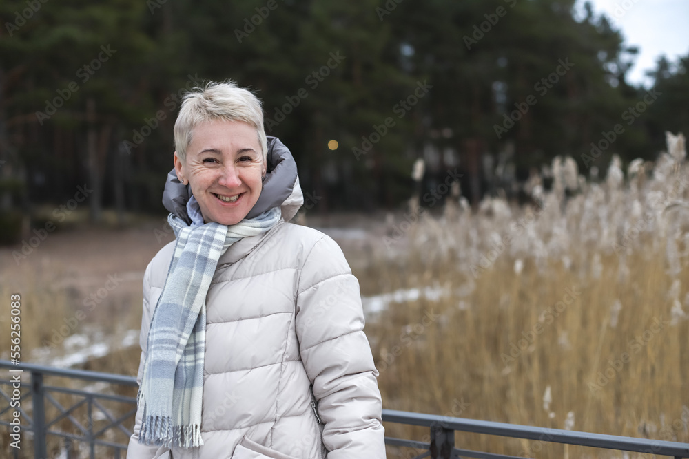 Fototapeta premium Portrait of happy beautiful elderly senior retired woman in age outdoors in forest or park at winter cold day, smiling and calm enjoy weather