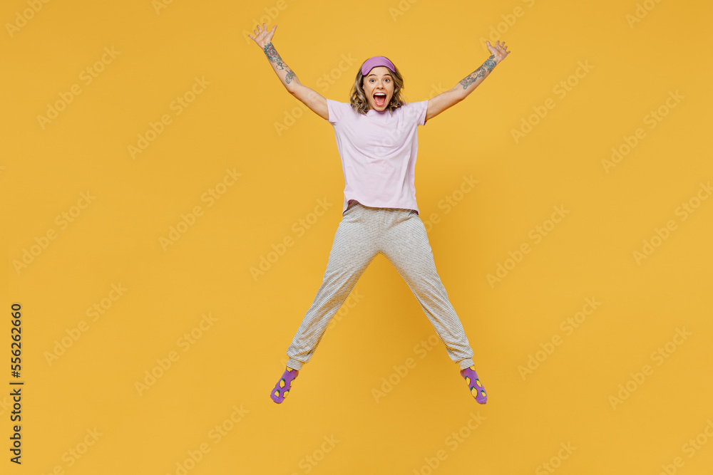 Full body fun young woman she wears purple pyjamas jam sleep eye mask rest relax at home jump high with outstretched hands legs isolated on plain yellow background studio portrait Night nap concept.