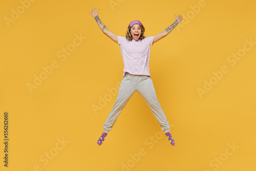 Full body fun young woman she wears purple pyjamas jam sleep eye mask rest relax at home jump high with outstretched hands legs isolated on plain yellow background studio portrait Night nap concept.