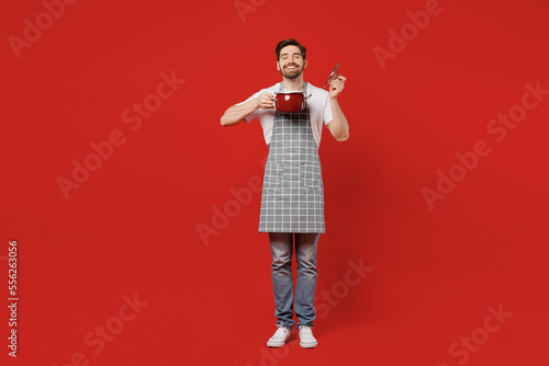 Full body young male housewife housekeeper chef cook baker man in grey apron holding in hand saucepan open pot lid enjoy smell close eyes isolated on plain red background studio Cooking food concept photo