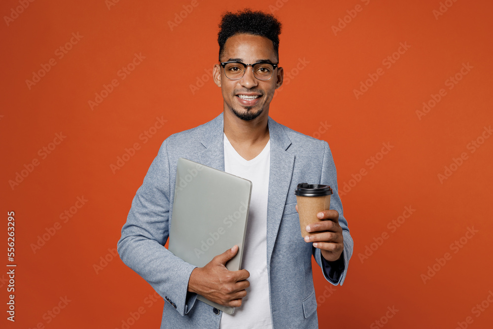 Young employee IT business man corporate lawyer wear classic formal grey suit shirt glasses work in office hold closed laptop pc computer drink coffee isolated on plain red orange background studio. Young employee IT business man corporate lawyer wear classic formal grey suit shirt glasses work in office hold closed laptop pc computer drink coffee isolated on plain red orange background studio.
