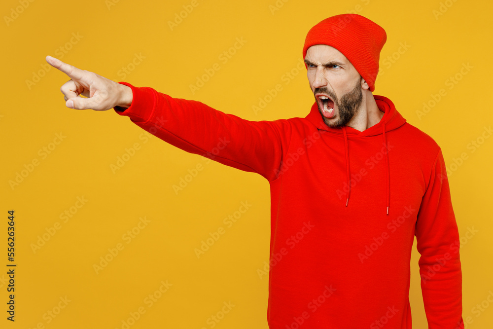 Young mad sad furious indignant angry strict caucasian man wears red hoody hat pointing index finger aside scream isolated on plain yellow color background studio portrait. People lifestyle concept. Young mad sad furious indignant angry strict caucasian man wears red hoody hat pointing index finger aside scream isolated on plain yellow color background studio portrait. People lifestyle concept.