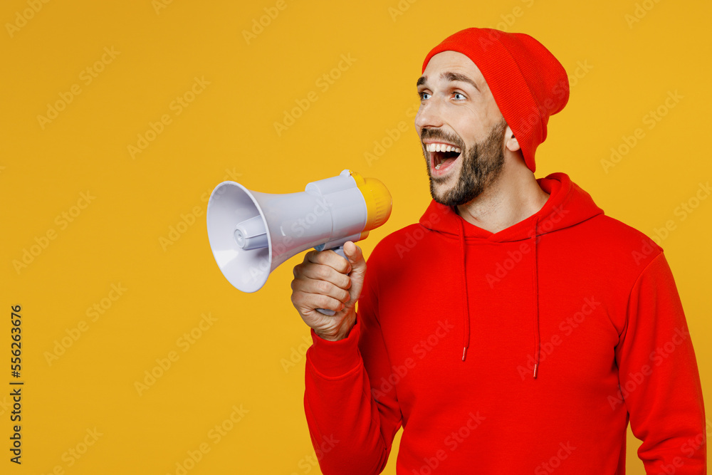 Surprised young shocked excited man wearing red hoody hat hold megaphone scream announces discounts sale Hurry up isolated on plain yellow color background studio portrait. People lifestyle concept Surprised young shocked excited man wearing red hoody hat hold megaphone scream announces discounts sale Hurry up isolated on plain yellow color background studio portrait. People lifestyle concept