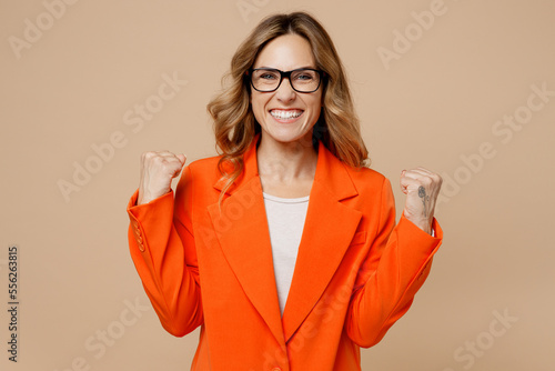 Young successful employee business woman corporate lawyer wear classic formal orange suit glasses work in office do winner gesture celebrate clenching fists isolated on plain beige background studio.