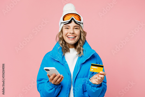 Snowboarder woman wear blue suit goggles mask hat ski jacket use mobile cell phone credit bank card book tour isolated on plain pastel pink background. Winter extreme sport hobby weekend trip concept.