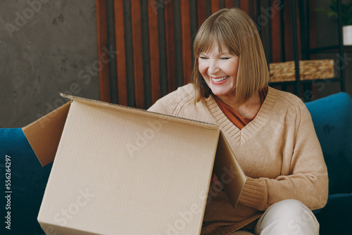 Elderly fun smiling woman 50s years old wears casual clothes sits on blue sofa look at cardboard box delivery parcel stay at home flat rest relax spend free spare time in living room indoor grey wall.
