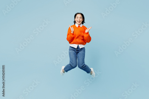 Full body exultant astonished young woman of Asian ethnicity wear orange sweater glasses jump high spread hands isolated on plain pastel light blue cyan background studio. People lifestyle concept.