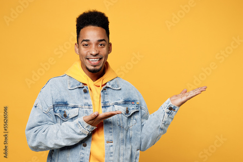 Young fun man of African American ethnicity wear denim jacket hoody point hands arms aside indicate on workspace area copy space mock up isolated on plain yellow background. People lifestyle concept.