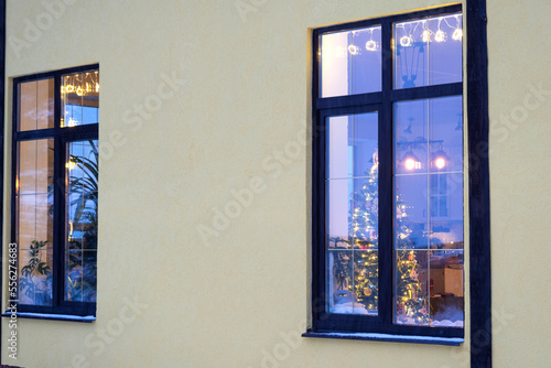 Cozy festive window of the house outside with the warm light of fairy lights garlands inside - celebrate Christmas and New Year in a warm home. Christmas tree, bokeh, snow on pine trees and snowfall