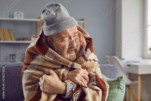 Senior man freezing at home in winter. Elderly man wearing hat and wrapped in blanket sitting on sofa in chilly room with low temperature, in house with bad broken thermostat, in very cold wintertime