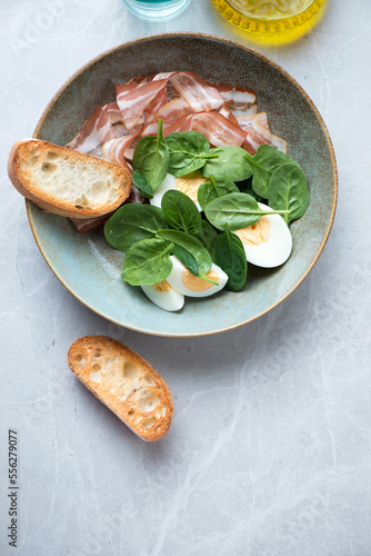 Plate with spinach, bacon, boiled chicken eggs and ciabatta, above view on a grey stone background, vertical shot with space