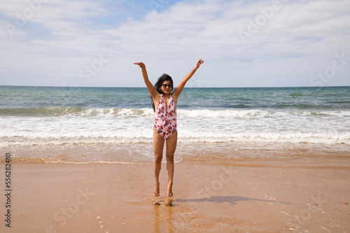 Beautiful and young latin woman is walking along the shore of the beach and posing for photos, while raising her arms and jumping as a symbol of freedom and well-being. Holiday and travel concept.