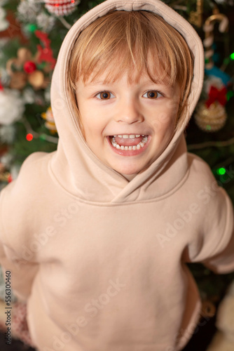 a happy blond boy in a light jacket with a hood on his head stands at the Christmas tree 