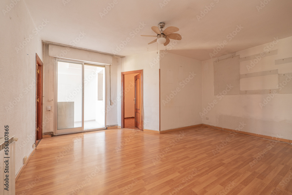 Empty living room with sapele wood interior doors with white painted walls, ceiling fan with wooden blades and enclosed terrace with aluminum and glass sliding doors Empty living room with sapele wood interior doors with white painted walls, ceiling fan with wooden blades and enclosed terrace with aluminum and glass sliding doors