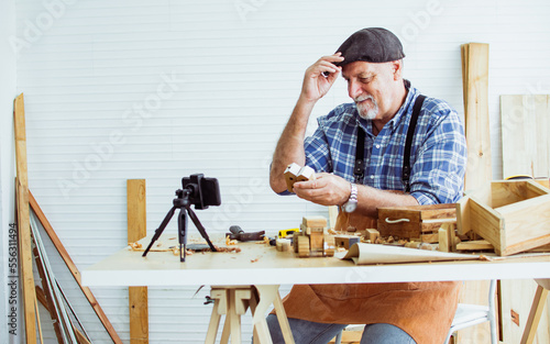 Senior old Caucasian man wearing check shirt, apron, making DIY wooden furniture, using mobile phone, streaming live video clip, selling product in social media at home. Retirement, Hobby Concept.