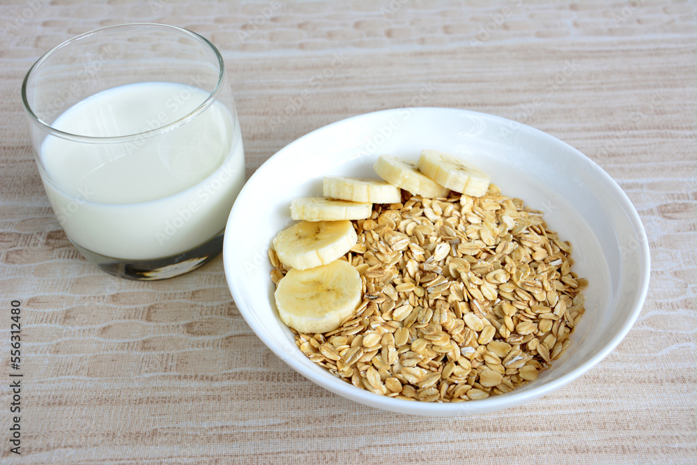oatmeal flakes in white bowl with banana slices and glass of milk isolated on pastel background