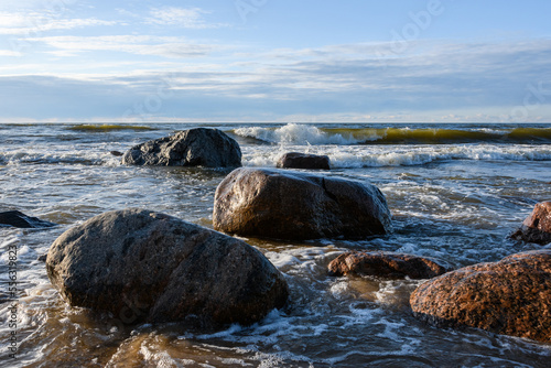 Sea waves roll and break on the coastal granite rocks. Sunny spring evening, Narva Bay, Baltic Sea