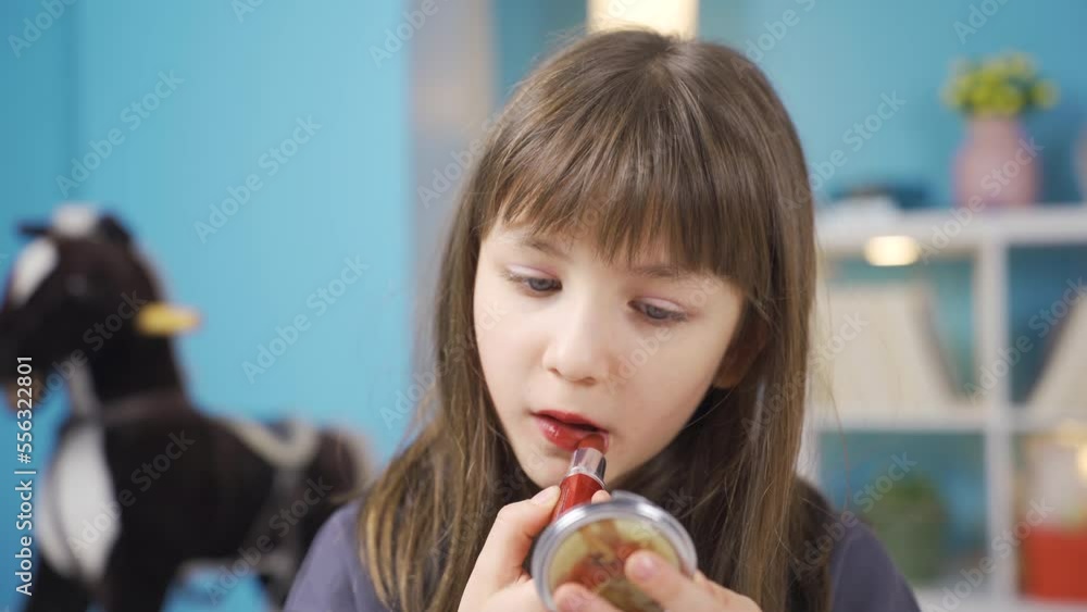 Little girl paints her lips with lipstick. Doing makeup.
Funny little girl having fun learning to do makeup in front of small mirror in her room.
