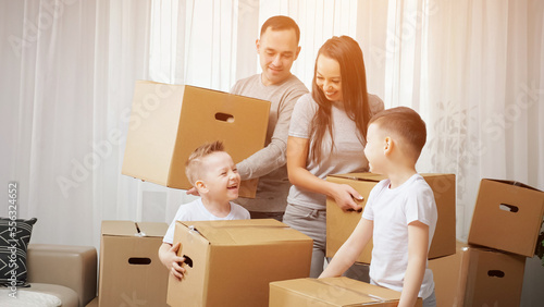 Family explores new apartment and gets ready to move carrying packages. Preschooler boy and junior schoolboy enjoy moving into new house, sunlight