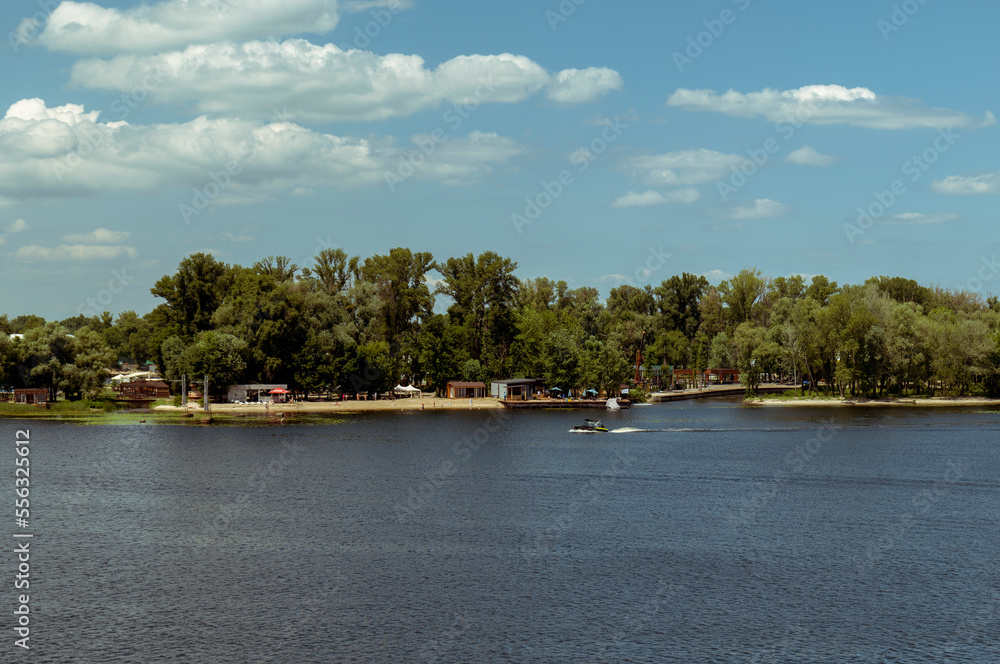 View across the water of the river to the beach recreation area near the forest. White clouds in the blue sky