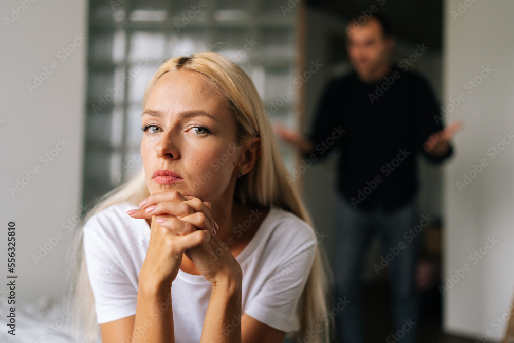 Portrait of tired young woman sadness looking at camera sitting on bed on background of aggressive husband shouting on girlfriend at home. Concept of family scandal, crisis, domestic violence, abuse. Portrait of tired young woman sadness looking at camera sitting on bed on background of aggressive husband shouting on girlfriend at home. Concept of family scandal, crisis, domestic violence, abuse.