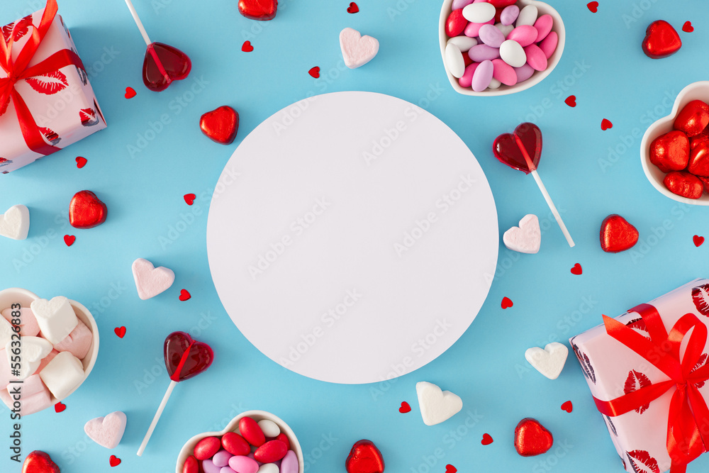 St Valentine's Day concept. Flat lay photo of red gift boxes, heart shaped saucers with sweets candies and lollipops, heart marshmallow on pastel blue background with round card note in the middle. St Valentine's Day concept. Flat lay photo of red gift boxes, heart shaped saucers with sweets candies and lollipops, heart marshmallow on pastel blue background with round card note in the middle.