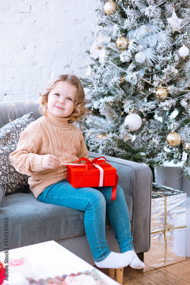 Naklejka premium cute happy little girl with curls in Santa hat is sitting on chair near Christmas tree with red gift box in her hands, New Year and Christmas family concept