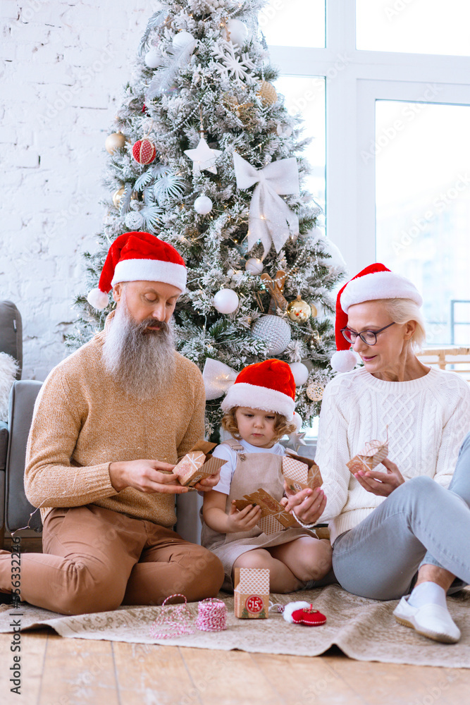 Fototapeta premium Christmas family grandmother, grandfather and granddaughter sit on sofa near Christmas tree and open advent calendar, pretty girl and active happy seniors give each other gift box