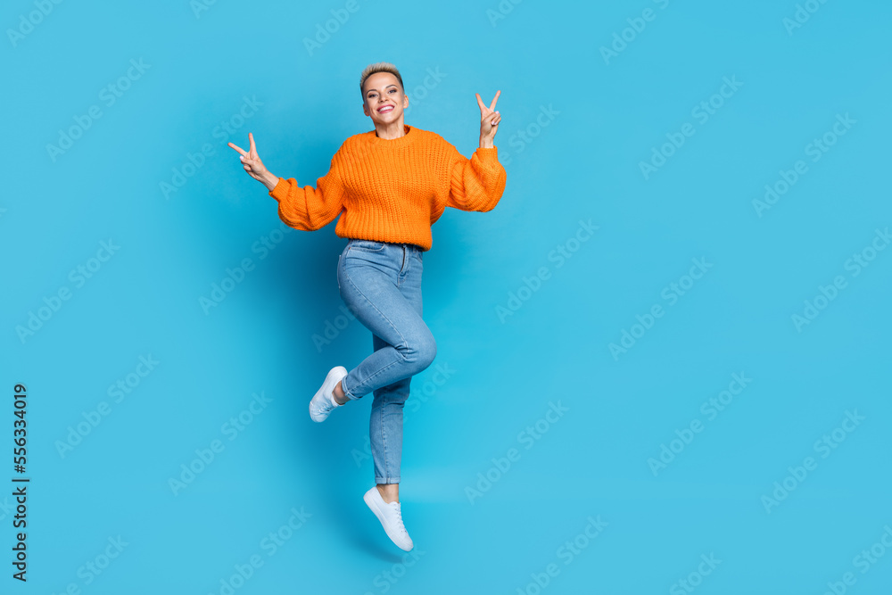 Fototapeta premium Full length photo of pretty sweet lady wear orange sweatshirt jumping high v-signs empty space isolated blue color background