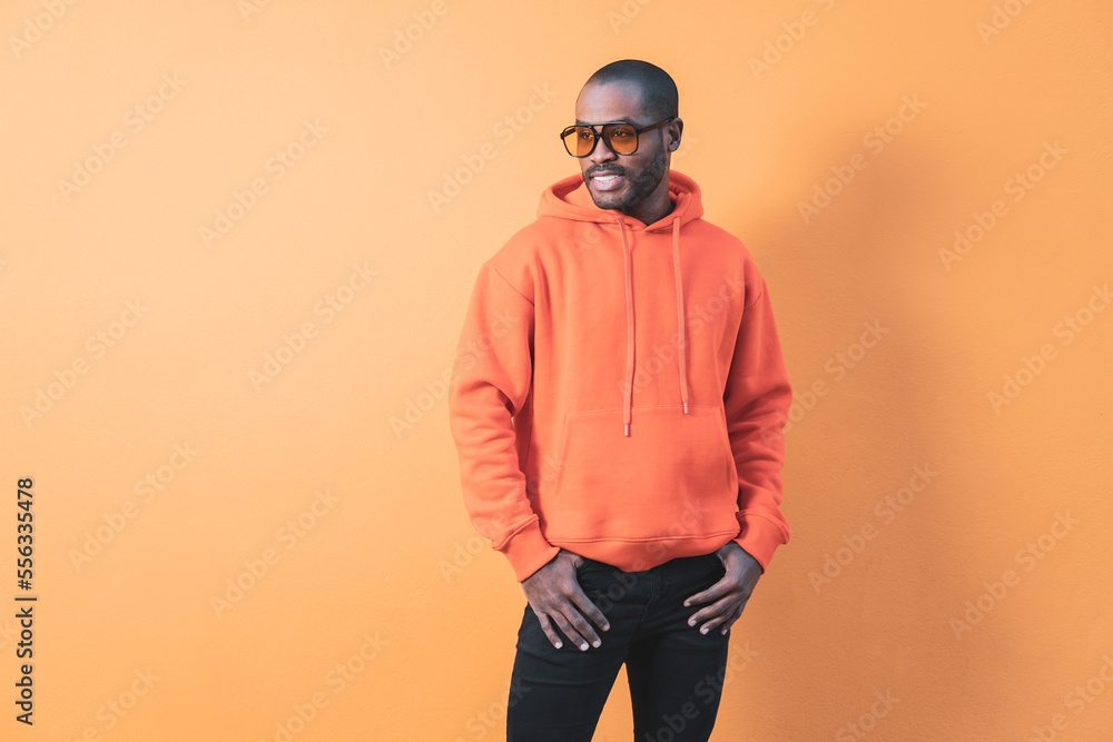 Fashion portrait of smiling handsome dark-skinned elegant man wearing hoodie with hat and jeans. Fashionable man posing near an orange wall in sunglasses