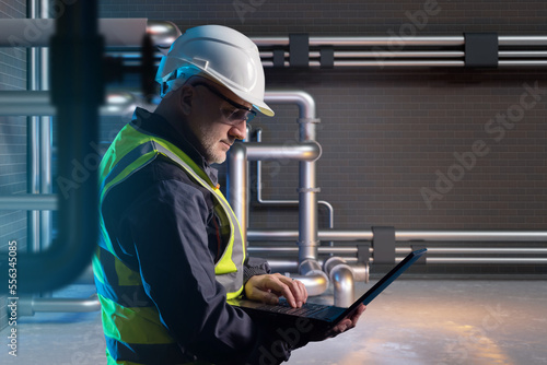 Man with laptop inside factory. Industrial room with metal pipes. Inspector supervises chemical enterprise. Man inspector in protective helmet. Industrial chemical factory employee.