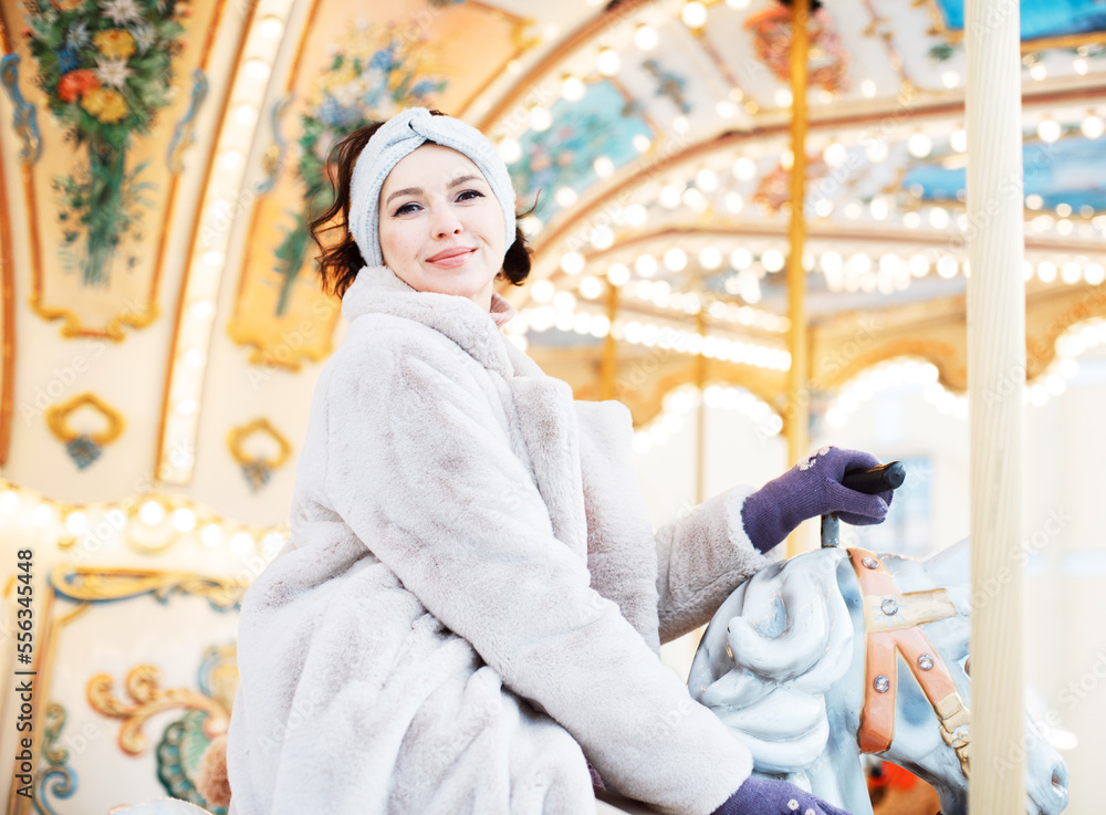 Beautiful and cheerful brunette woman in a fur coat rides on a carousel