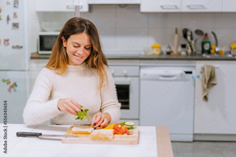 Vegetarian woman cooking a vegetable sandwich in the kitchen at home. Placing the arugula Vegetarian woman cooking a vegetable sandwich in the kitchen at home. Placing the arugula