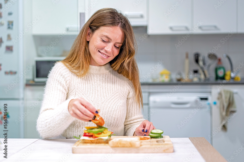 Vegetarian woman cooking a vegetable sandwich in the kitchen at home. Preparing it by putting tomato Vegetarian woman cooking a vegetable sandwich in the kitchen at home. Preparing it by putting tomato