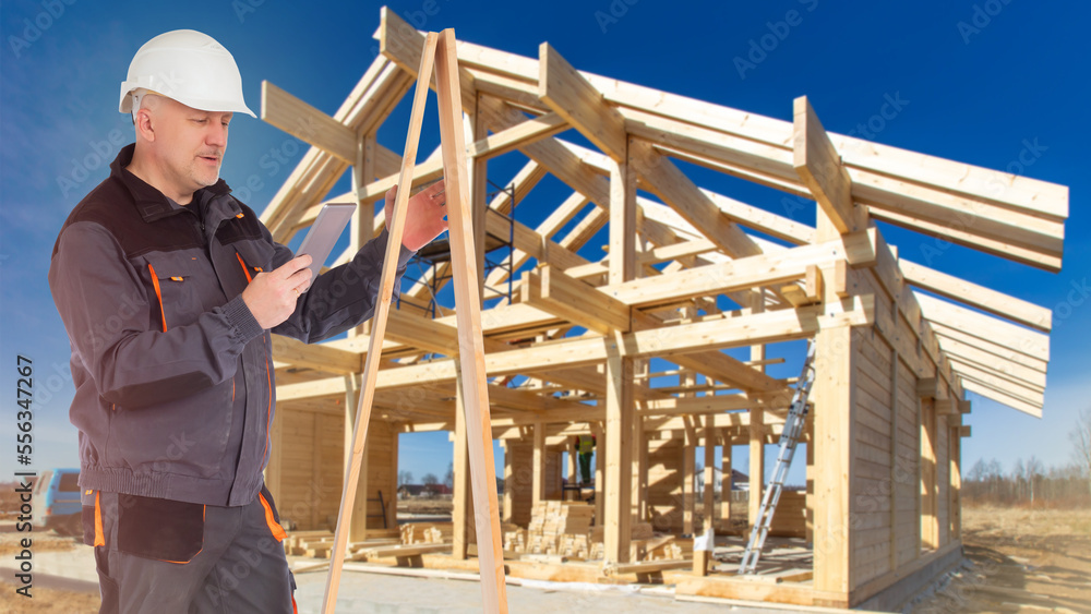 Man builder. Construction wooden house. Guy with tablet is building house. Construction frame cottage. Man in gray uniform builder. Worker using app for builders. Brigadier with planks under blue sky Man builder. Construction wooden house. Guy with tablet is building house. Construction frame cottage. Man in gray uniform builder. Worker using app for builders. Brigadier with planks under blue sky