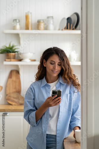 Young pleasant smiling Latin woman housewife standing in cozy kitchen holding smartphone  watching cooking video recipe. Happy European female spending time at home on mobile phone. Gadget addiction