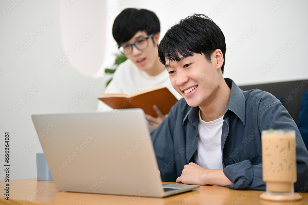 Two of smart young Asian male college students looking at laptop screen, doing a project together Two of smart young Asian male college students looking at laptop screen, doing a project together