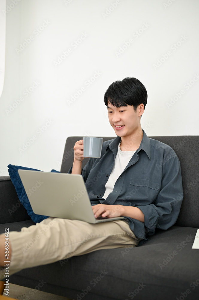 Portrait, Relaxed Asian man remote working from home, sipping coffee, using laptop on sofa Portrait, Relaxed Asian man remote working from home, sipping coffee, using laptop on sofa