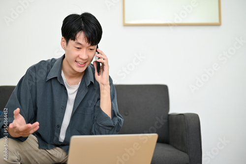 Happy young Asian man looking at laptop screen, talking on the phone with his colleagues