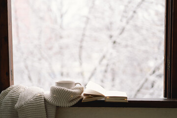 Cozy winter still life. Cup of hot tea and an open book with a warm sweater on a vintage wooden windowsill. Cozy home concept. Sweet home.