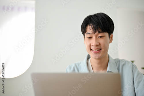 Happy cute young Asian man working on his business tasks on laptop computer at home.