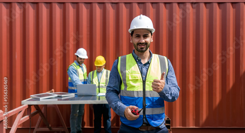 Project manager raise finger thumb up with a smile while holding walkie talkie. The team is meeting in the background with laptop computer and solar cell panel. Everyone wear safety vest and hardhat.