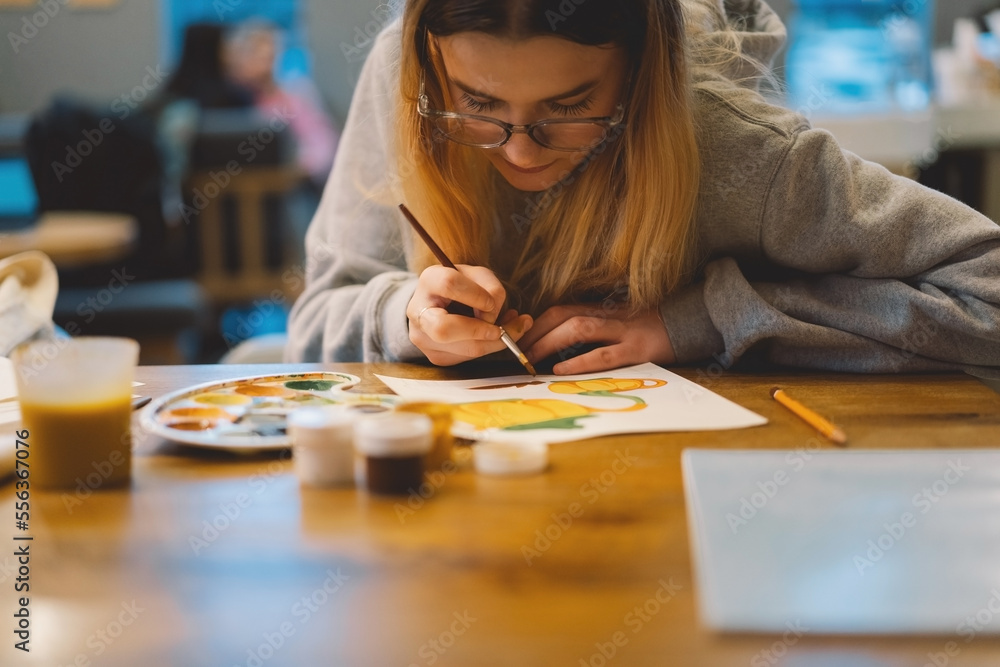 Fototapeta premium Pretty teenage girl draws a picture with poster paint. Front view of a drawing of a girl with a palette in her hand. A smiling young teenage girl draws a picture