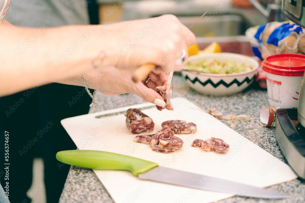 Young, latina, hispanic woman chopping salami on the kitchen counter with a bowl full of guacamole in the background.