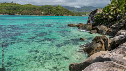 Corals are visible through the clear turquoise water of the ocean. Picturesque granite boulders and palm trees on the shore. In the distance is an island overgrown with tropical vegetation. Seychelles