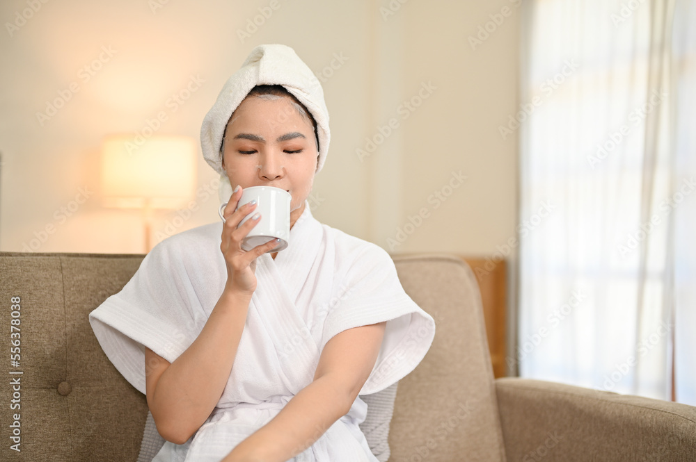 Relaxed Asian woman in bathrobe sipping morning coffee on the sofa in her living room. Relaxed Asian woman in bathrobe sipping morning coffee on the sofa in her living room.