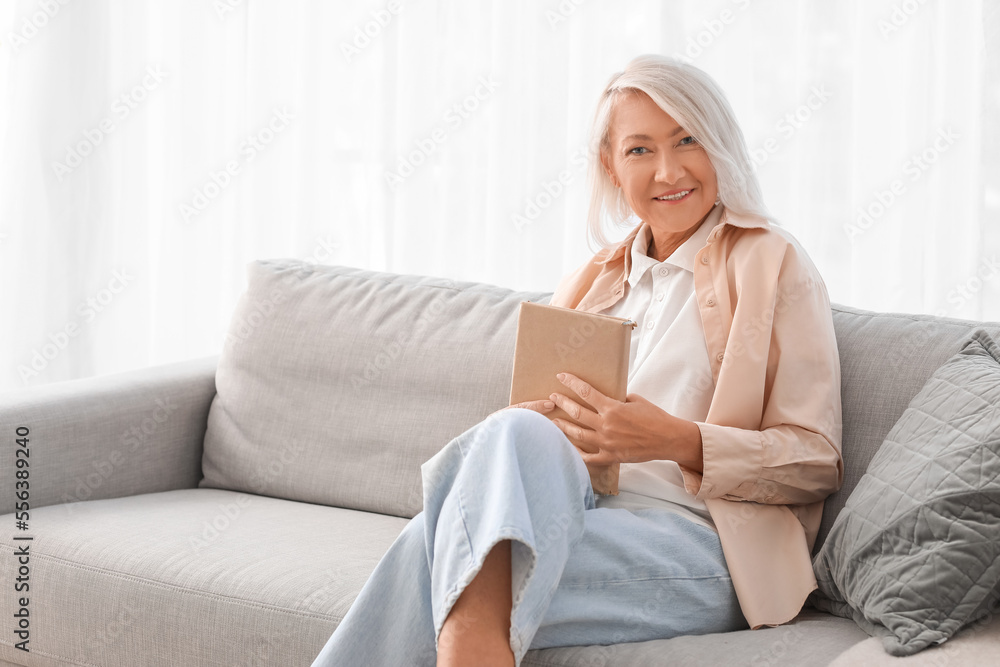 Mature woman with book sitting on grey sofa at home Mature woman with book sitting on grey sofa at home