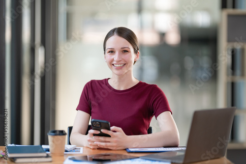 Attractive businesswoman sitting at her desk, holding a smartphone, smiling, and looking at the camera.