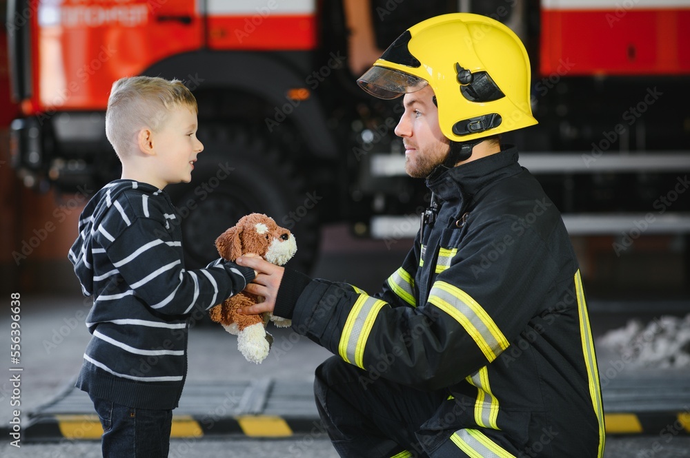 Obraz premium Portrait of rescued little boy with firefighter man standing near fire truck. Firefighter in fire fighting operation.
