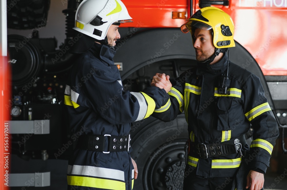 Naklejka premium Two firefighters in protective clothing in helmets with fire engine, friendly handshake